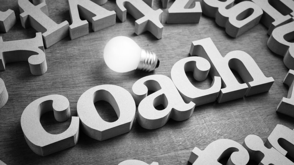 Wooden block letters spelling out 'coach' with a glowing light bulb placed above, symbolizing guidance and inspiration.